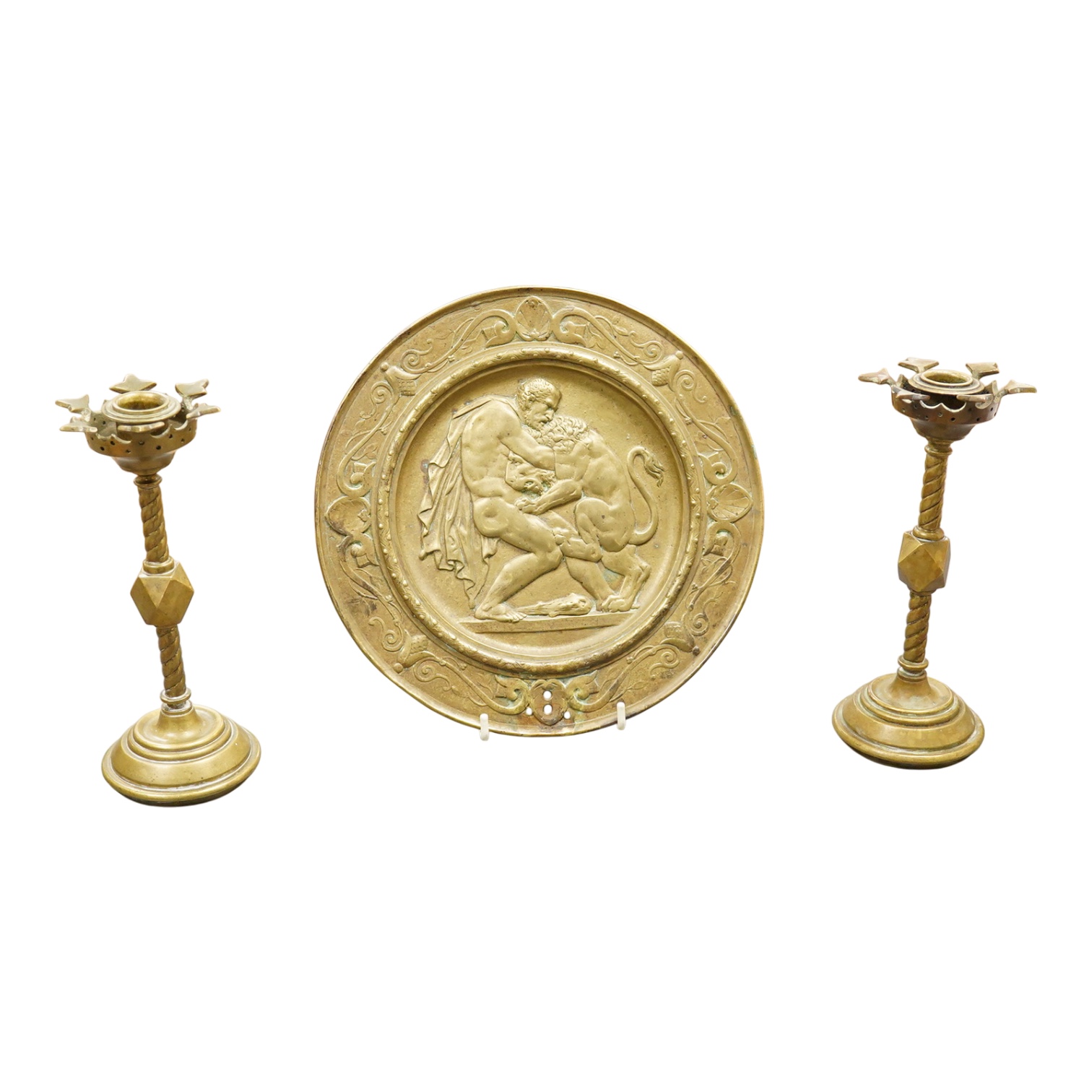 A pair of Pugin style ‘gothic’ brass candlesticks - 21cm high and a circular brass disc depicting Hercules and The Nemean Lion - 25cm in diameter